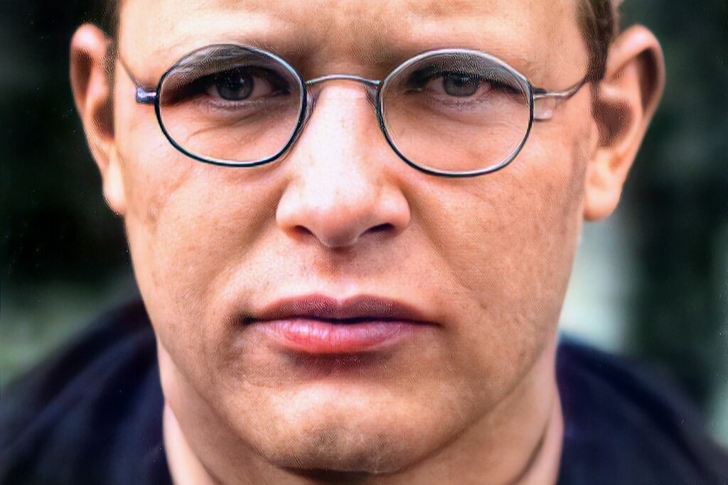 A close up of a 1930s man wearing wire-rimmed glasses, looking pensive.