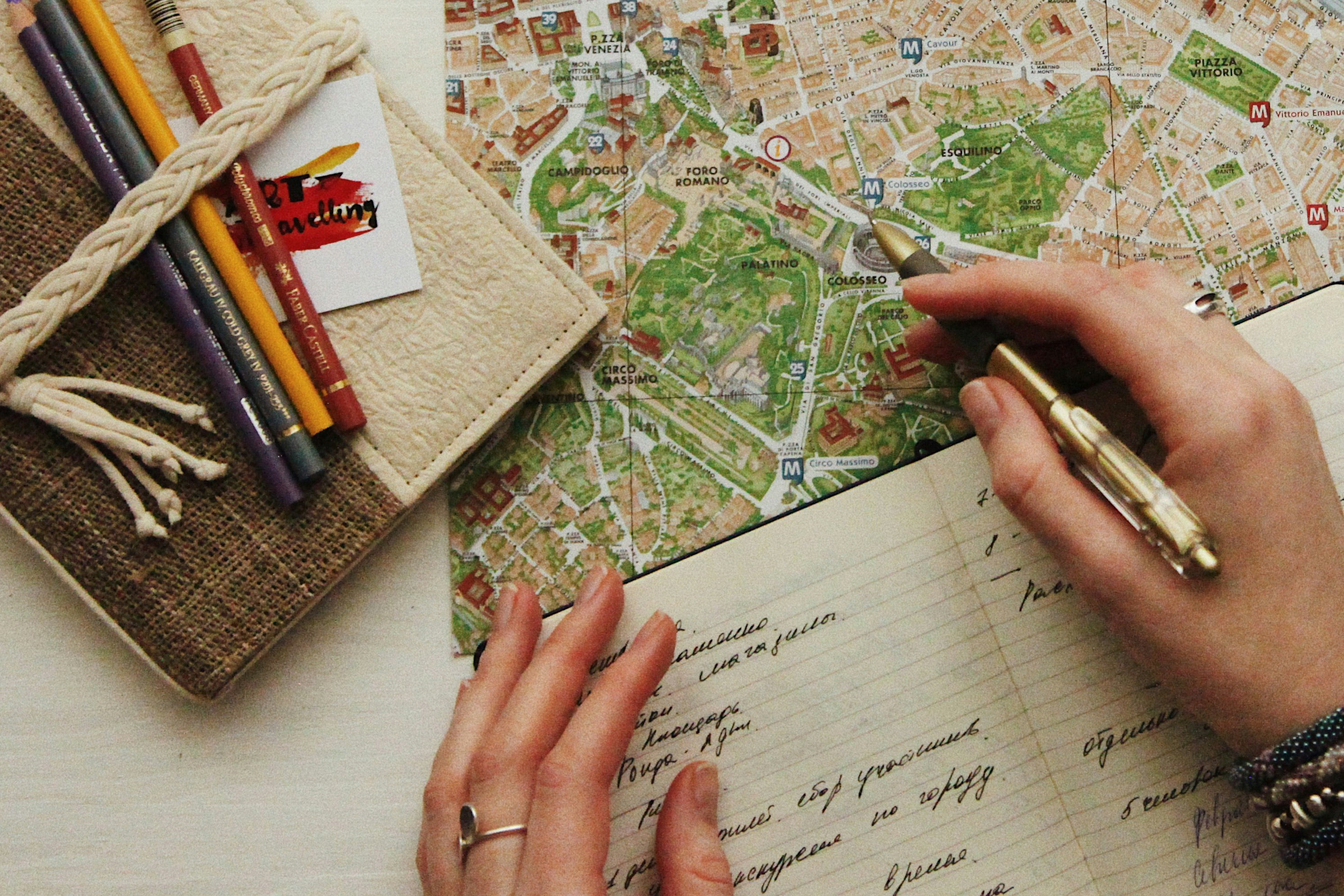 A hand holds a pen over a map, at the side is closed journal and colour pencils.