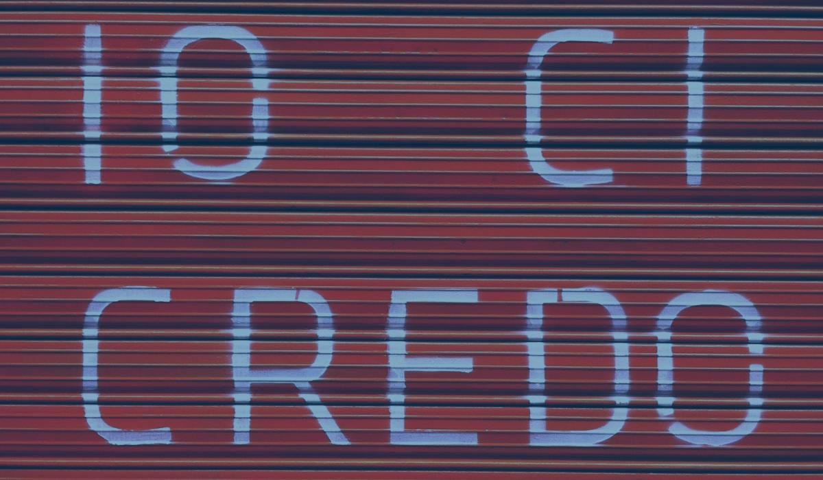 A red shutter door bears a large painted message reading 'io ci credo'