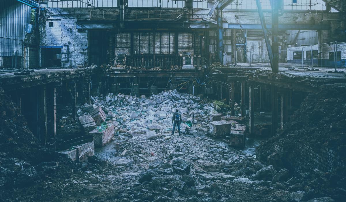 A photographer, standing next to a tripod, atop a pile of rubble is a destroyed factory.