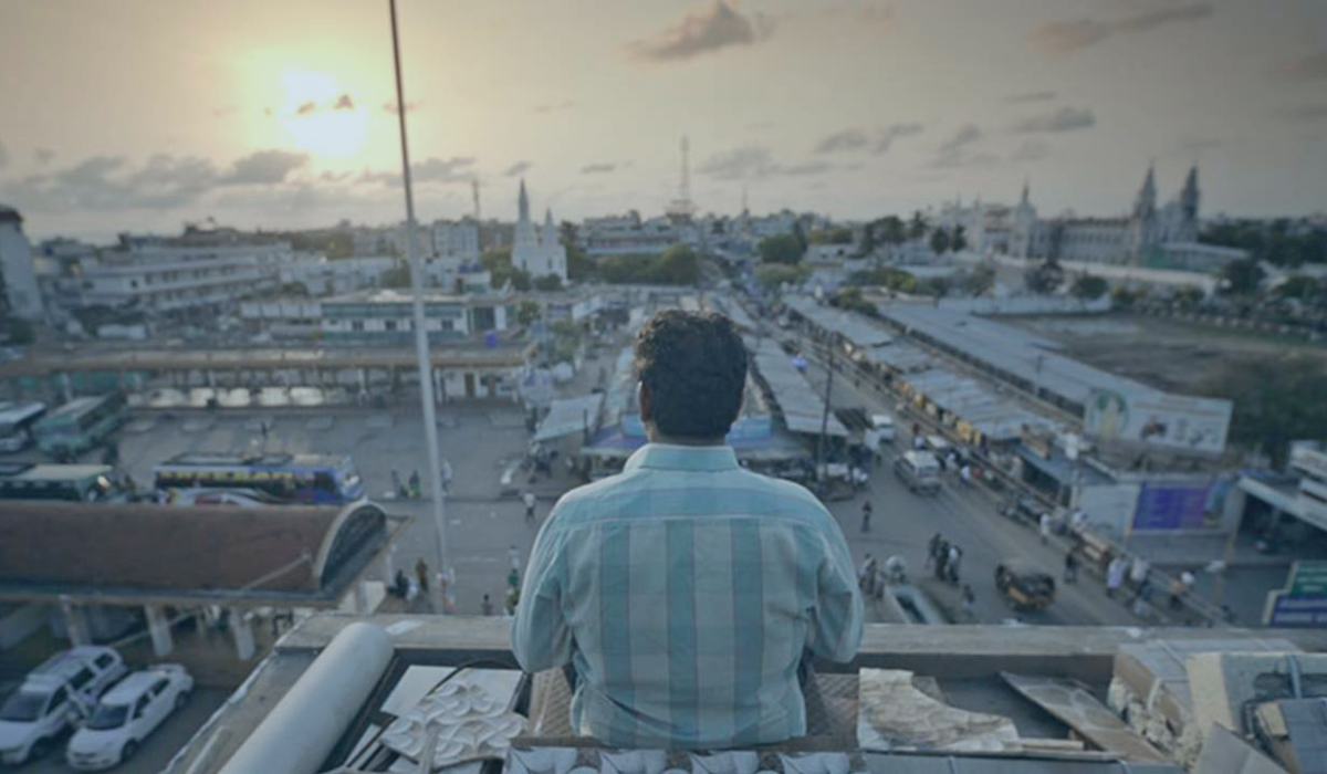 A man sits on the top of a building and looks out over the view of an Indian city as the sun sets.