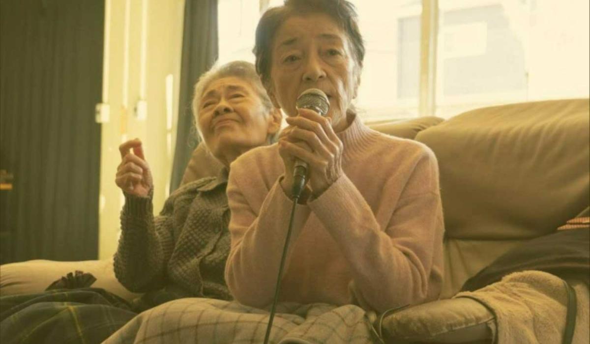 In a retirement home, a older person sings karaoke while the person behind waves a hand.