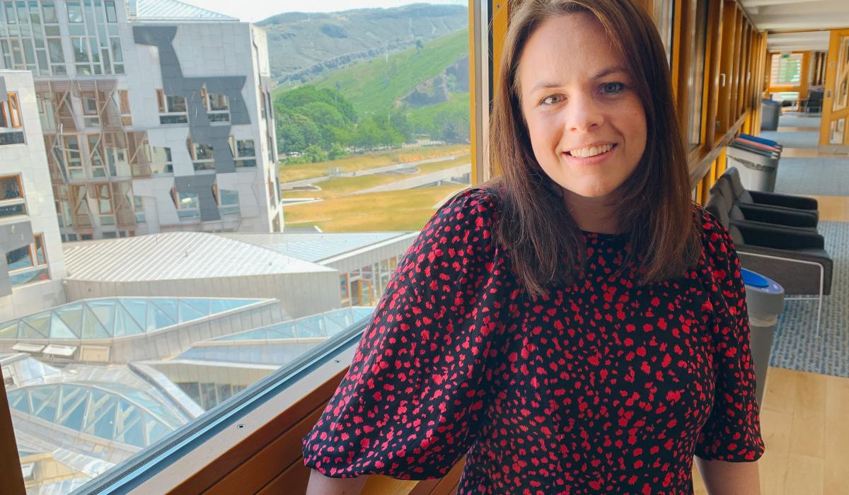 A women stands beside in a corridor beside a large window through which a wing of a building and a distant hillside can be see,