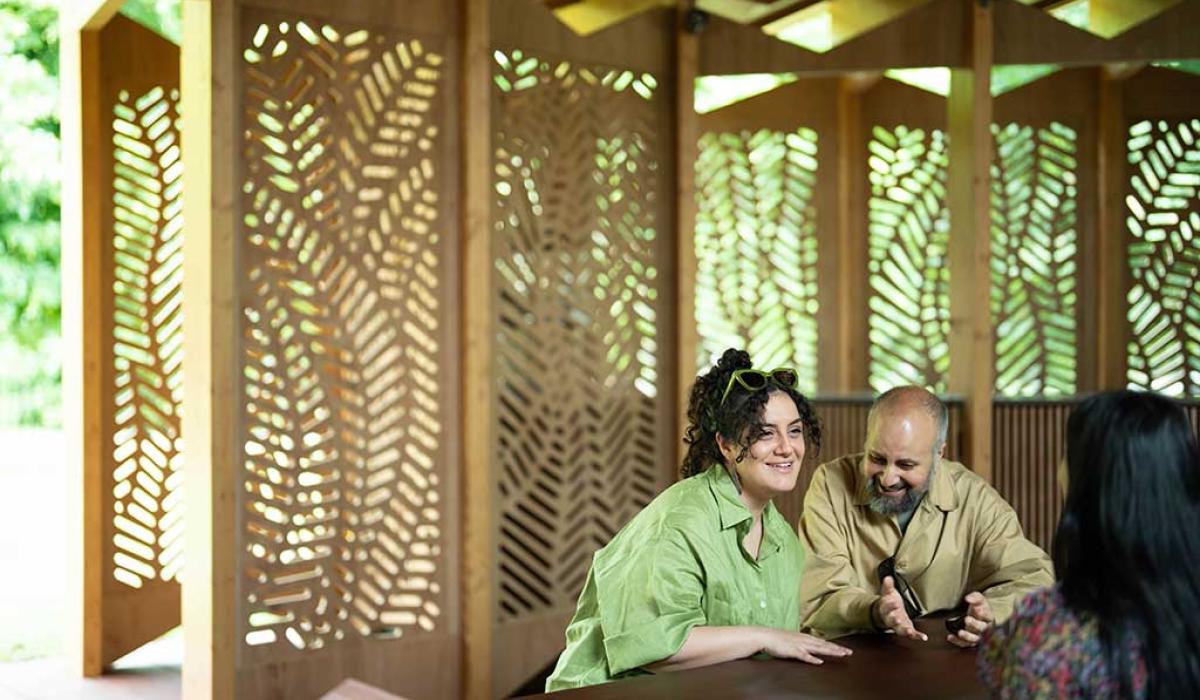 Around a table, against a backdrop of fret-cut wood, three people talk and listen to each other with great interest.