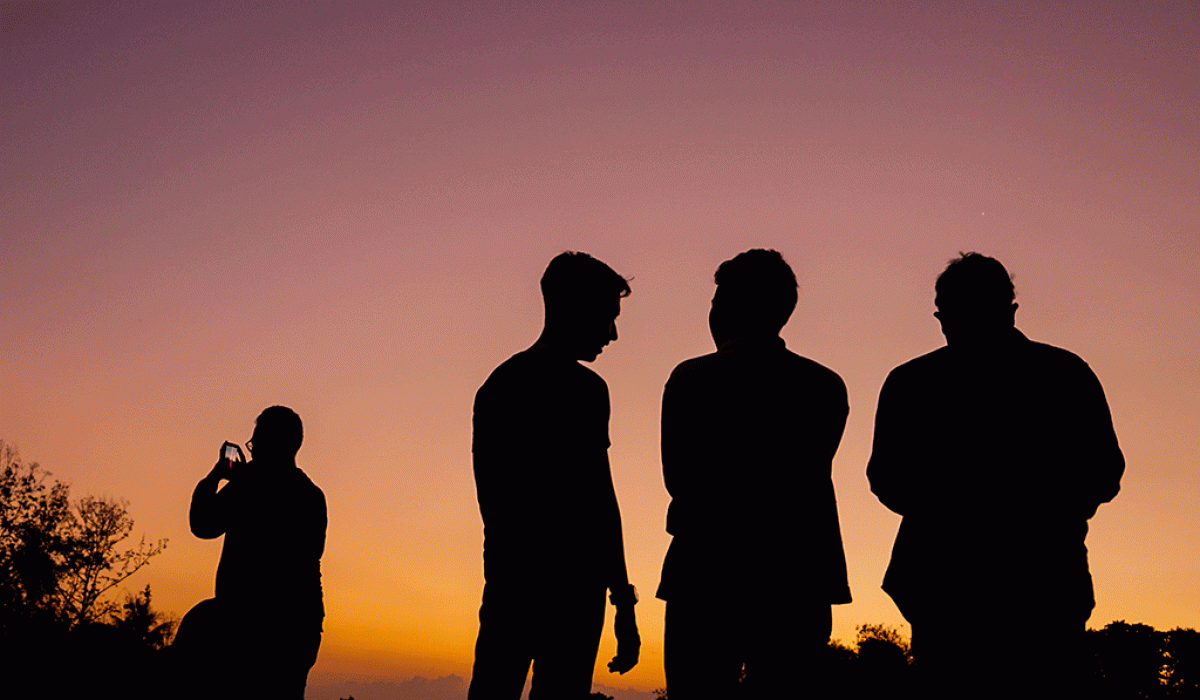 Four men stand silhoutted against a sunset, One stands apart on their phone.