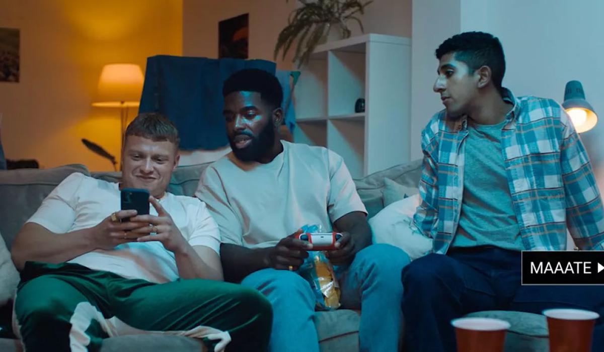 Three young men sit on a couch. One is leering at a phone while the others look on hesitantly