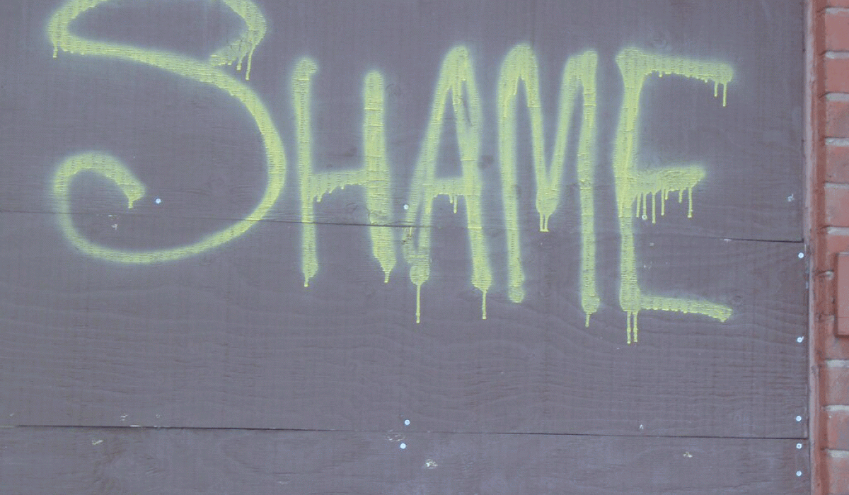 The word 'SHAME' spray painted onto a grey hoarding in lime green paint.