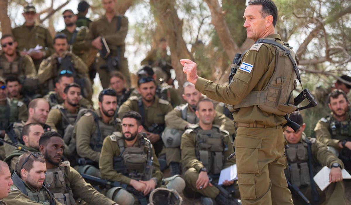 An army general in green fatigues stands and address a group of soldiers sitting, squatting and standing around him