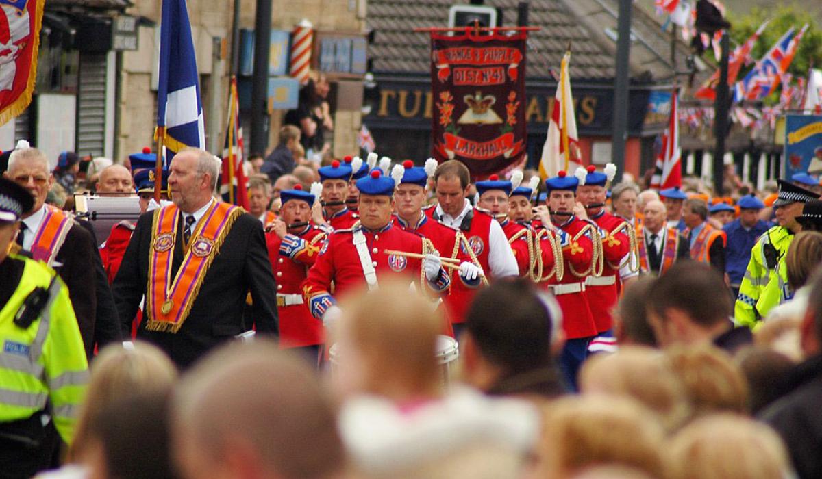 Across the heads of a roadside crowd, men wearing orange sashes and military band uniforms march along.