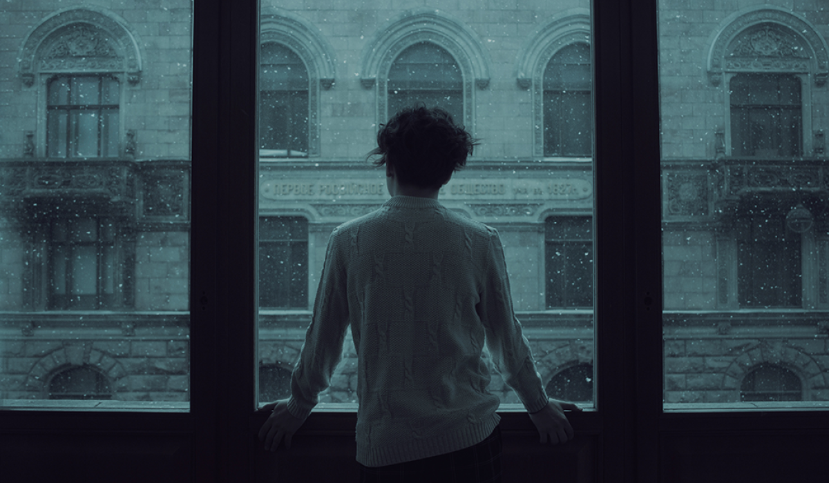 A person stand looking out a wide set of windows covered in rain.