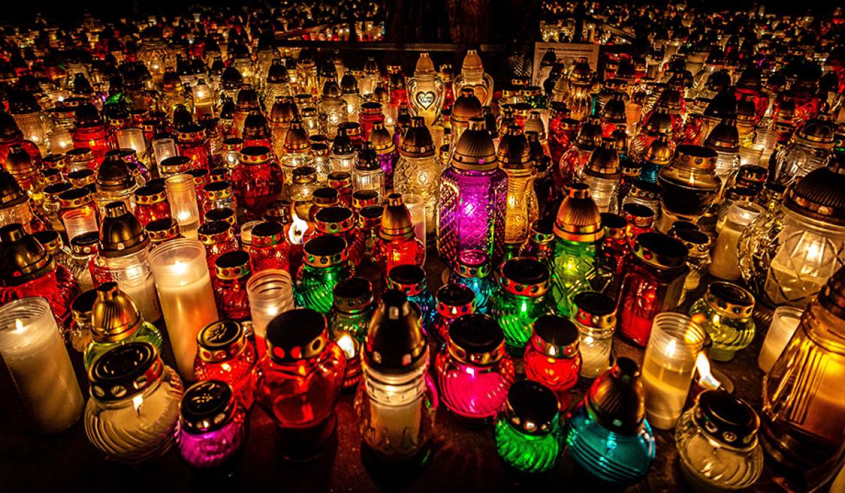 Dozens of candles in cloured jars and holders litter the ground of a cemetry.