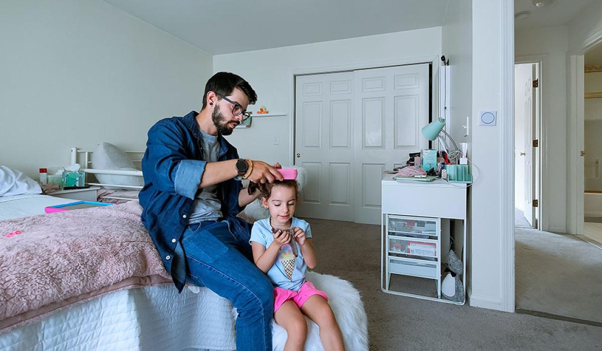 A father sits on a bed and fixes the hair of his daughter standing in front of him