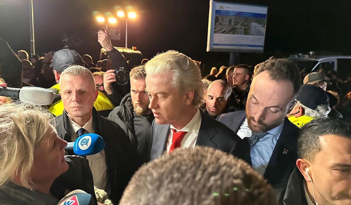 A politician in a suit stands amid a scrum of reporters holding microphones