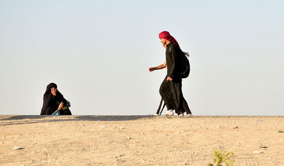 A black clad pilgrim squats and holds his head on the side of a desert road as other pilgrims walk along.