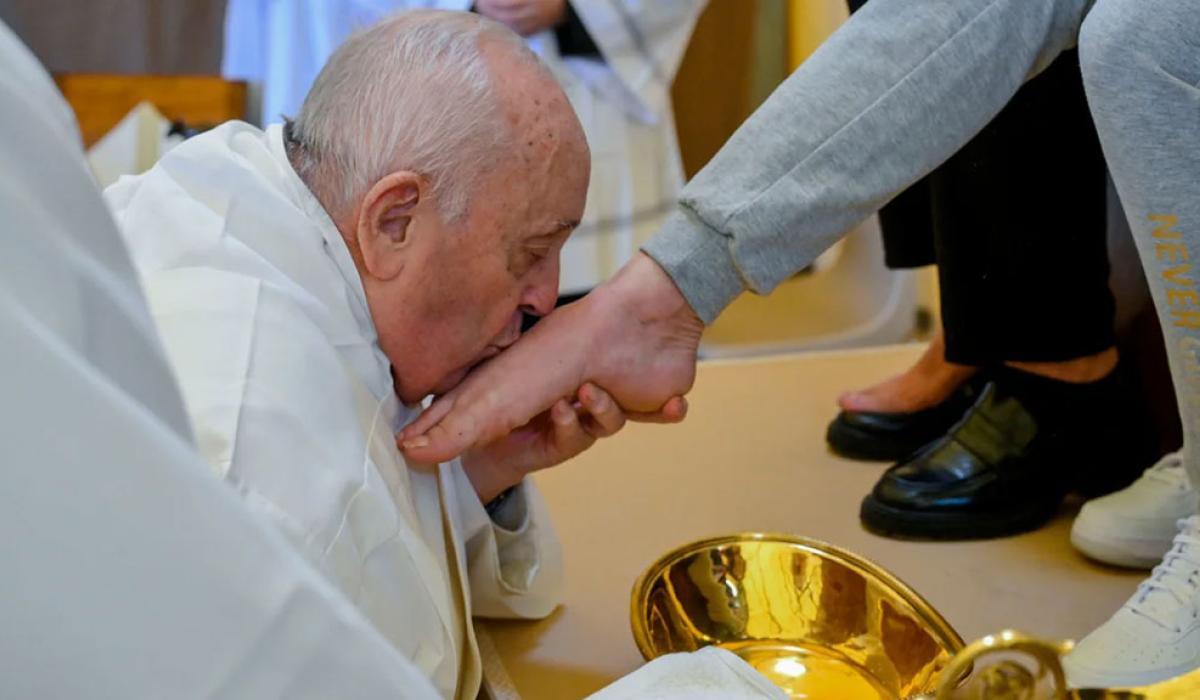 The Pope, wearing white, kneels, crades a bare foot, and kisses it.