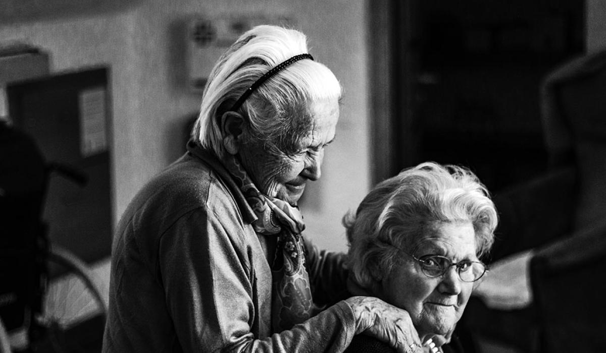 A elderly women in a care home stands and places her hands on the shoulders of a seated woman.