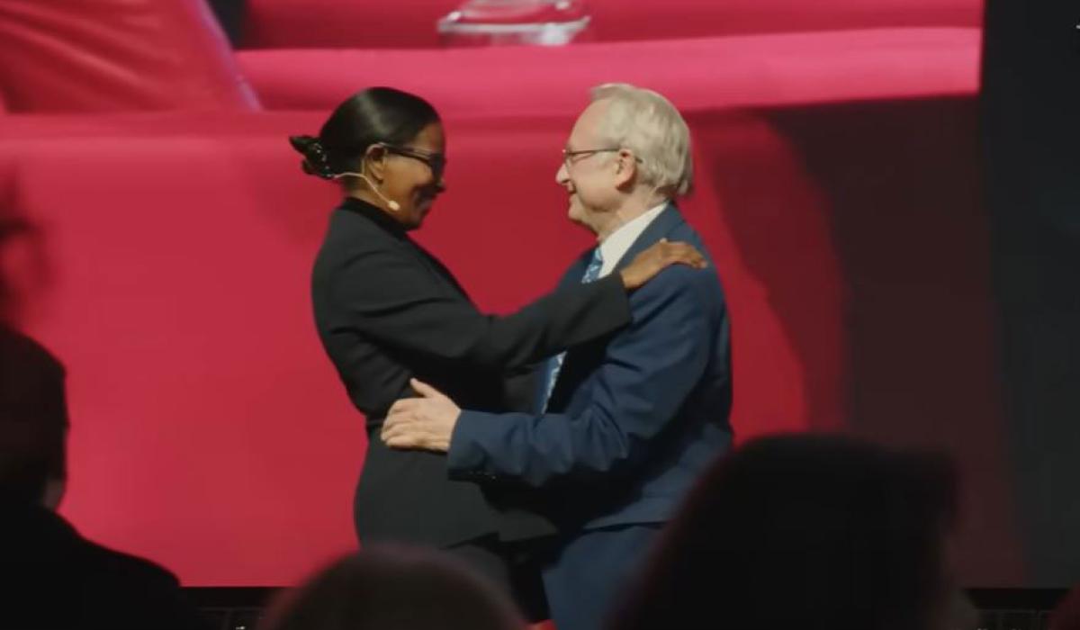 A man and woman speaker on a stage greet and embrace each other.