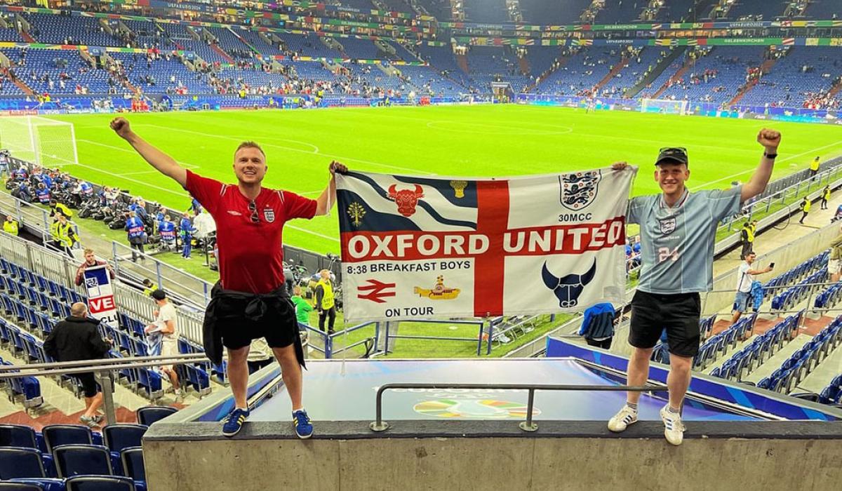 Two England fans stand in stadium seeting holding up their flag.