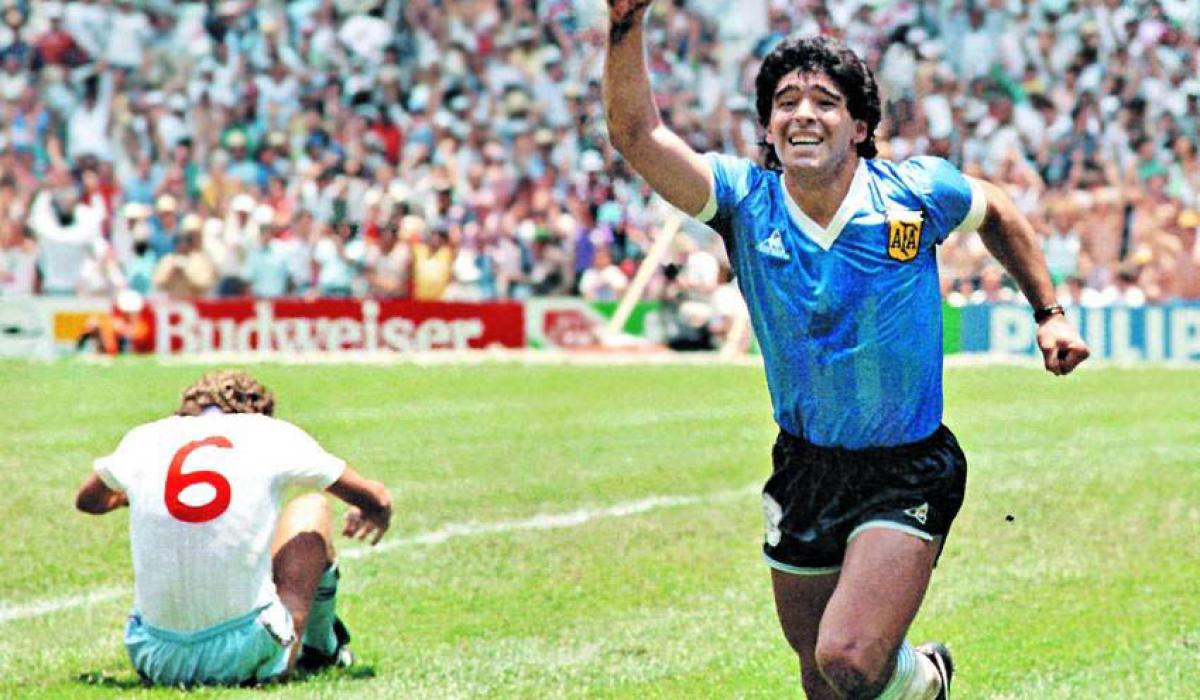 Maradona runs in celebration, holding a hand aloft as an England player sits dejected on the ground.