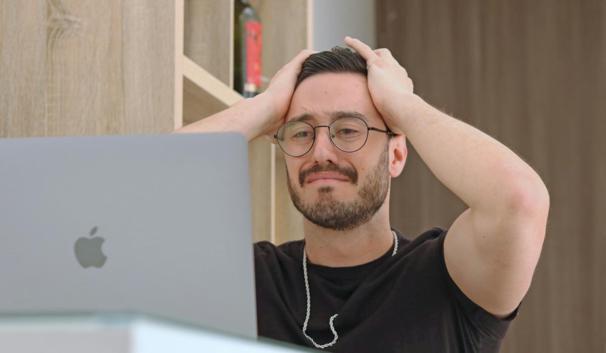 A man staring at a laptop grimmaces and holds his hands to his head.