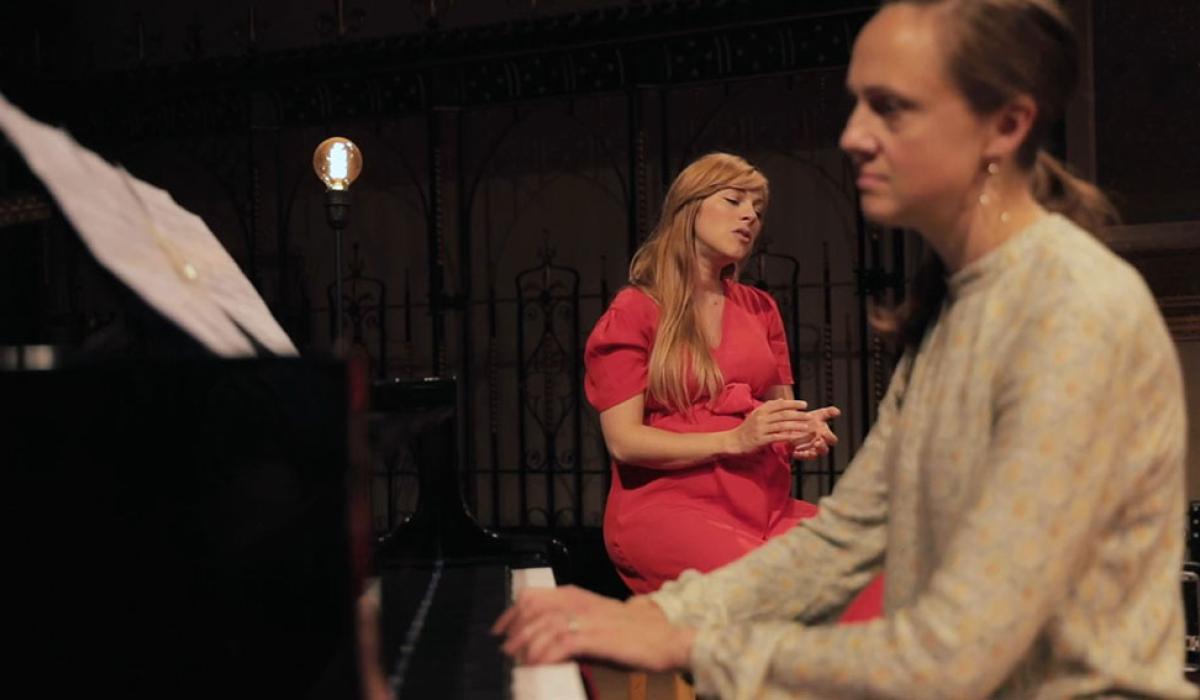 A pianist plays in the foreground and a seated singer gestures with eyes closed behind 