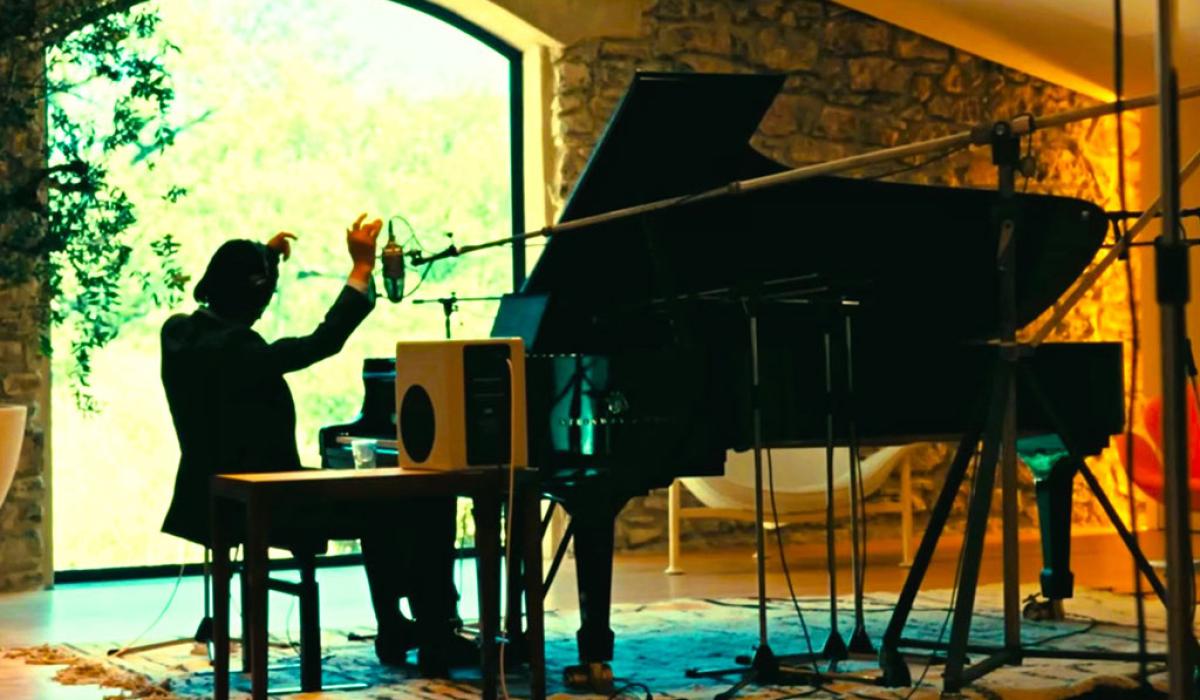 A pianist raises his arms while sitting at a grand piano amid recording equipment.