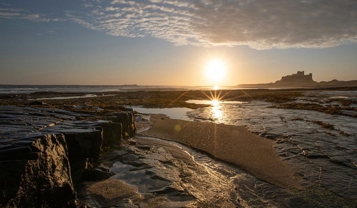 A rockpool on a beach reflects the sun, a castle stands beyond the sand dunes