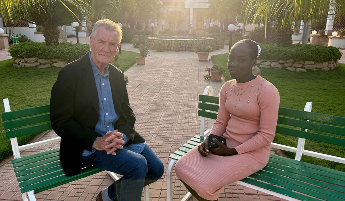 A TV interviewer sits across from a young woman in an outdoor setting