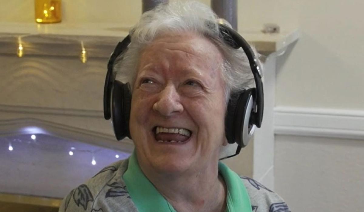 An elderly woman wearing headphone looks up and to the side with a big smile.