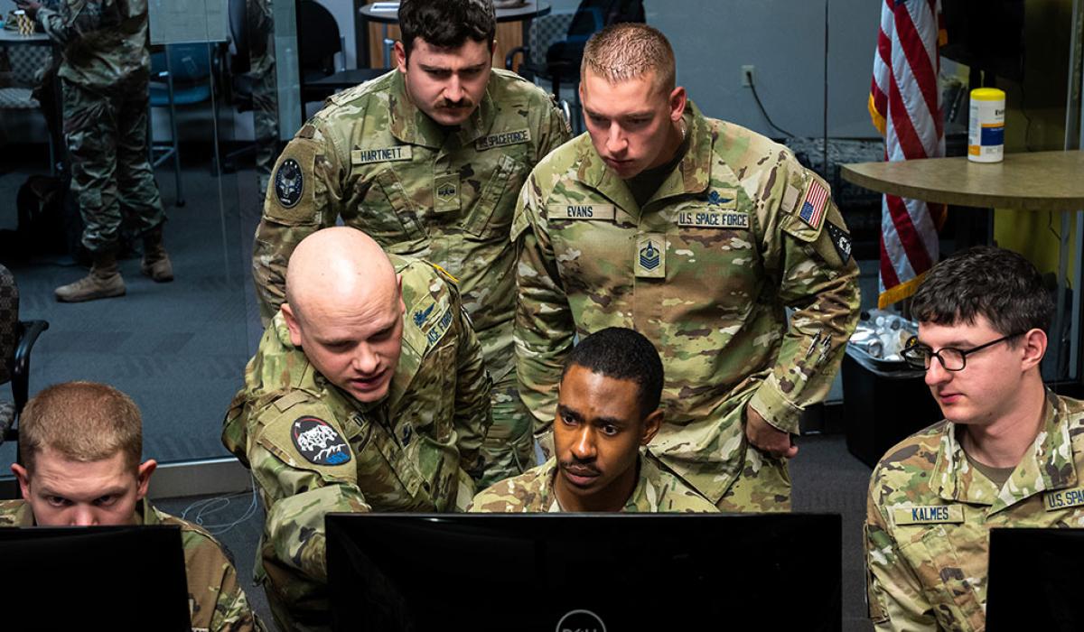 Military personnel, wearing camoflague uniform crowd round a computer monitor.