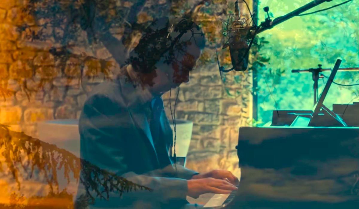 A pianist raises his arms while sitting at a grand piano amid recording equipment.