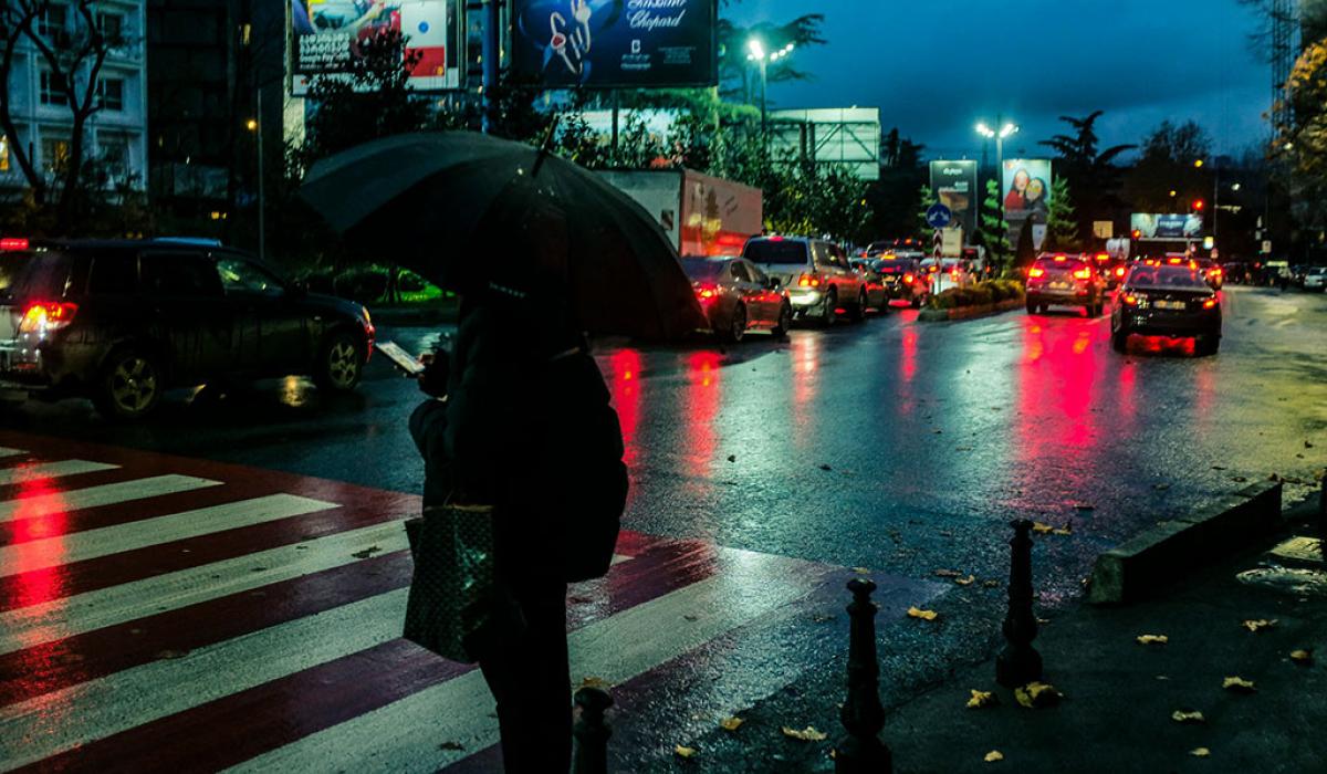 On a rainy night a pedestrian, holding a brolly, waits to cross a road.