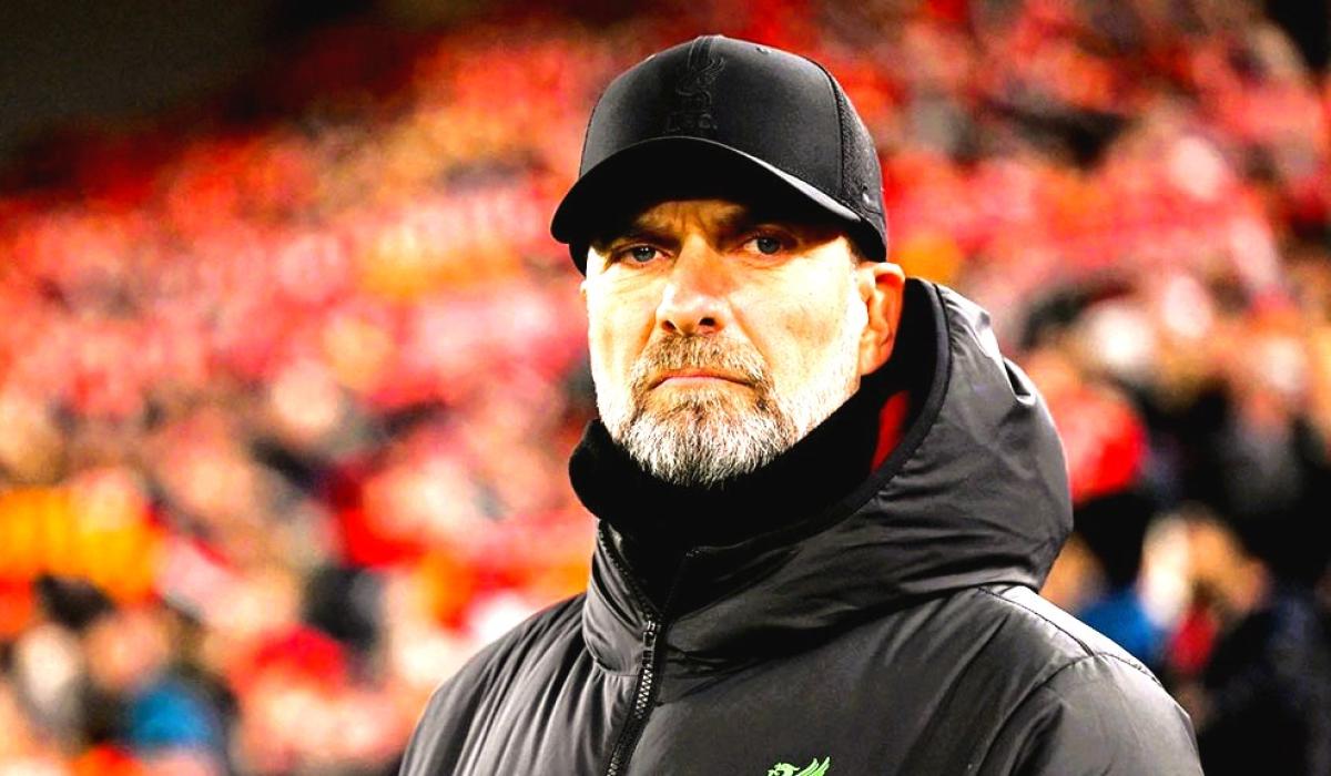 A stern looking  football manager stares hard, with a stand of supporters behind him.