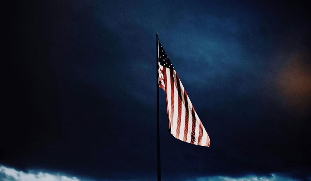 A US flag flutters under a dark brooding cloudy sky.