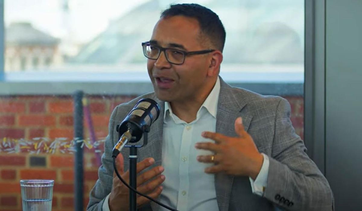 A man sitting at a table with a mic on it, raises his hands in gesture as he speaks. 