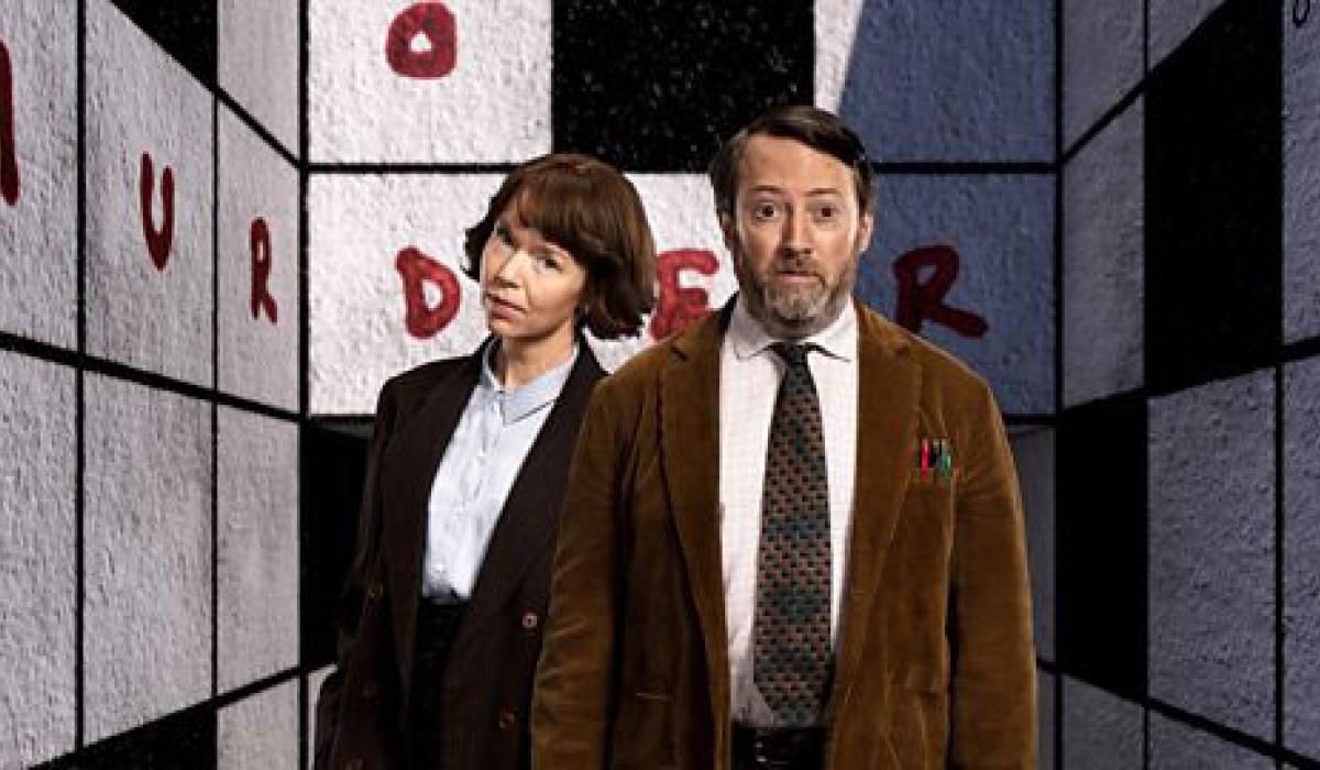 Two TV characters, a man and a woman, stand in front of a crossword cover walls of a room. 