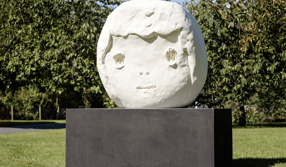 A large outdoor sculpture of a round child-like head sits on a dark plinth.