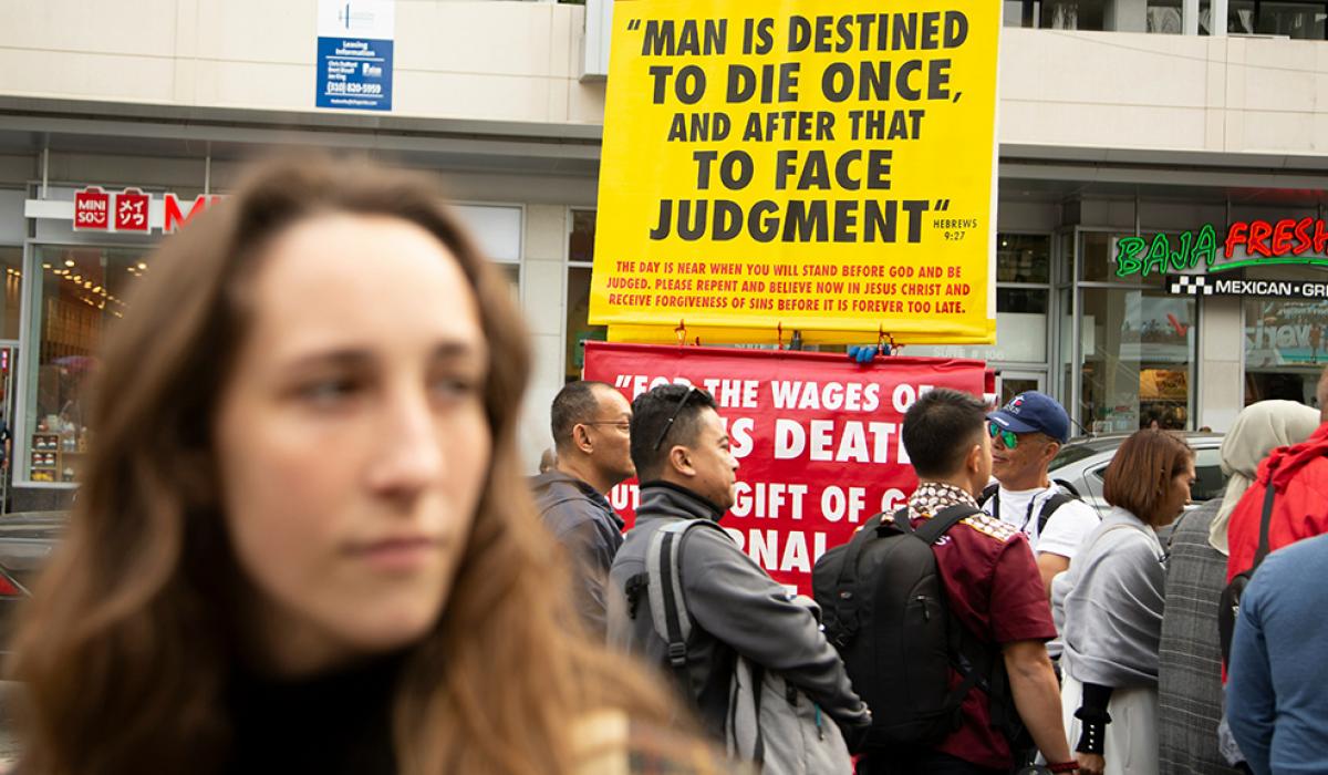 Behind a passer by a street peacher holds up a large yellow sign with a message on it.