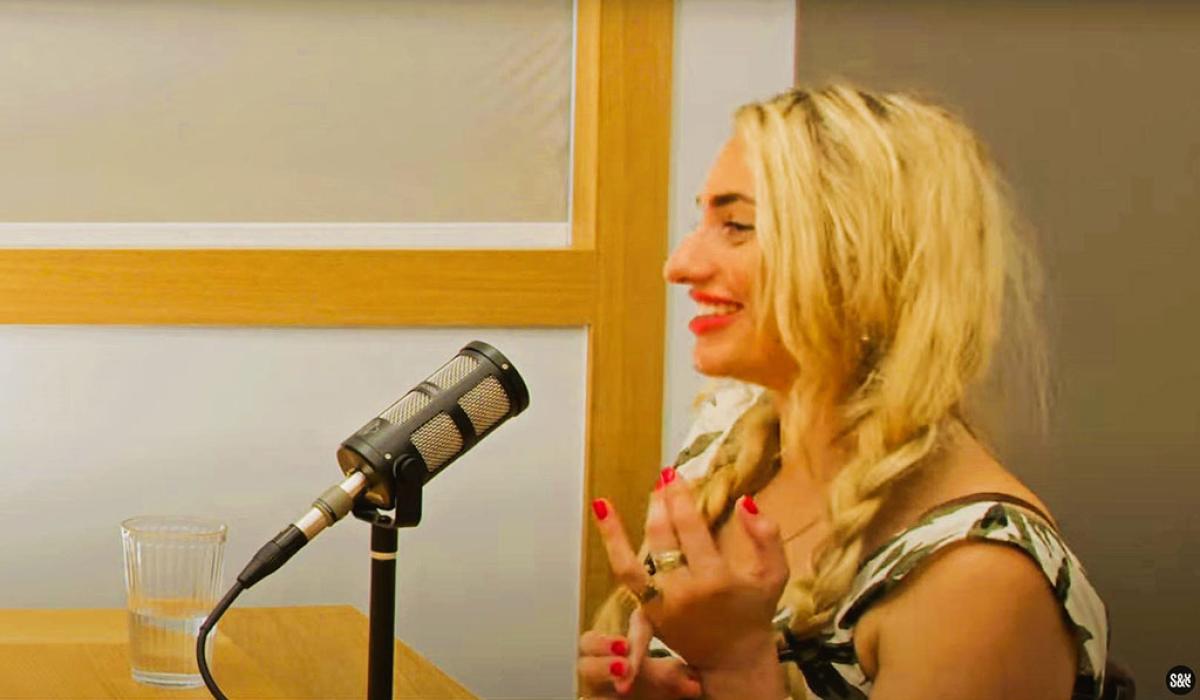  In profile a woman speaks into a microphone and holds her hands up from a desk.