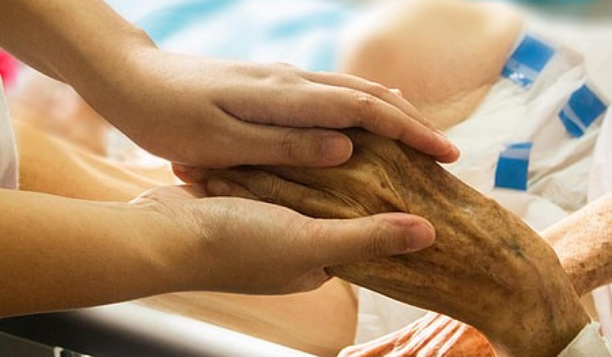 A younger hand holds a wrinkled older hand of someone in a bed.