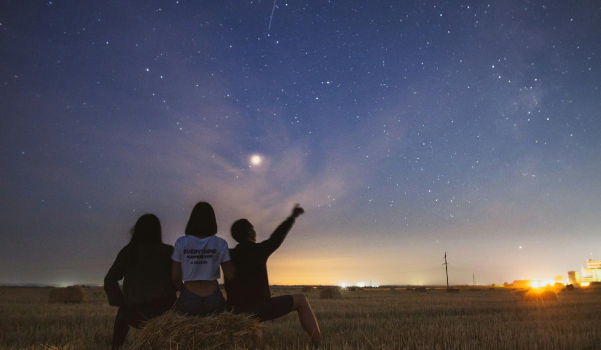 At dusk, three people sit on a field edge and look at the stars emerging.