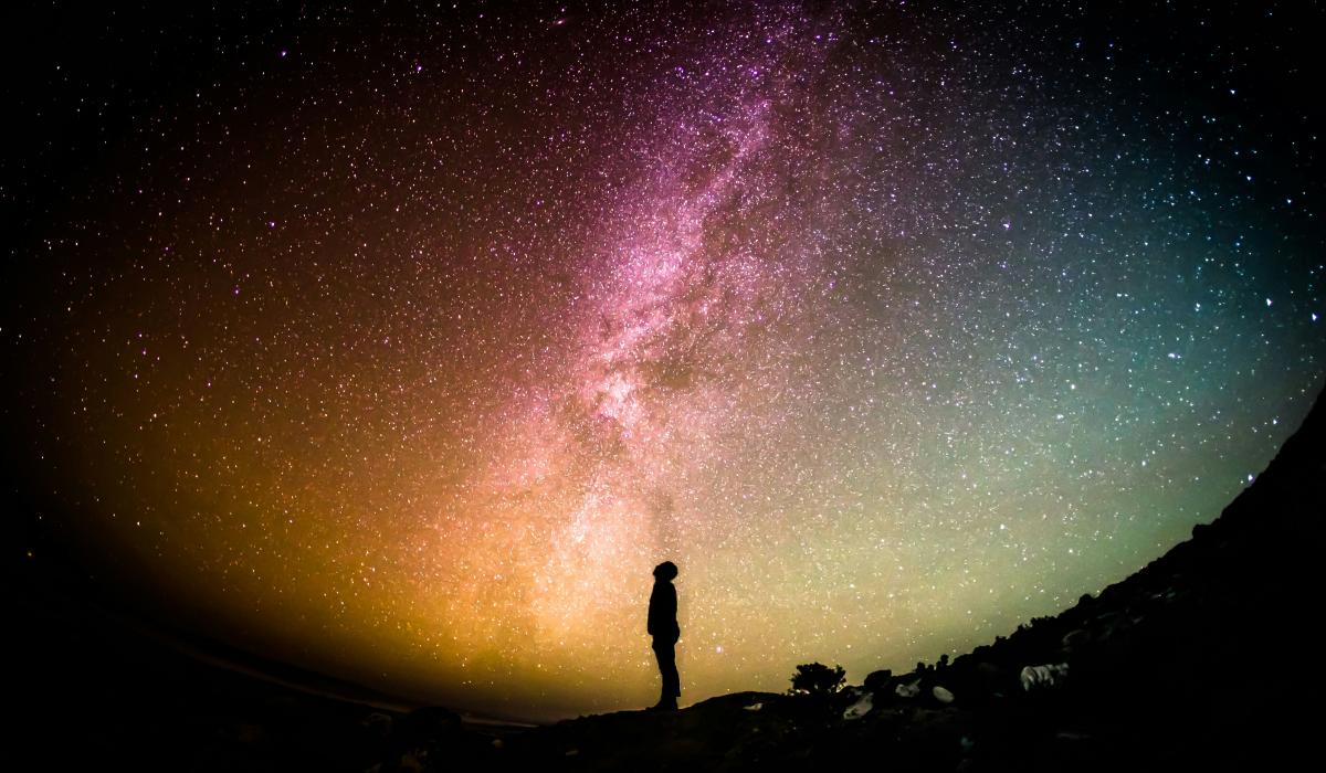 A fish eye lens view of a person standing silhouetted, looking up to a colourful night sky with the Milky Way across it