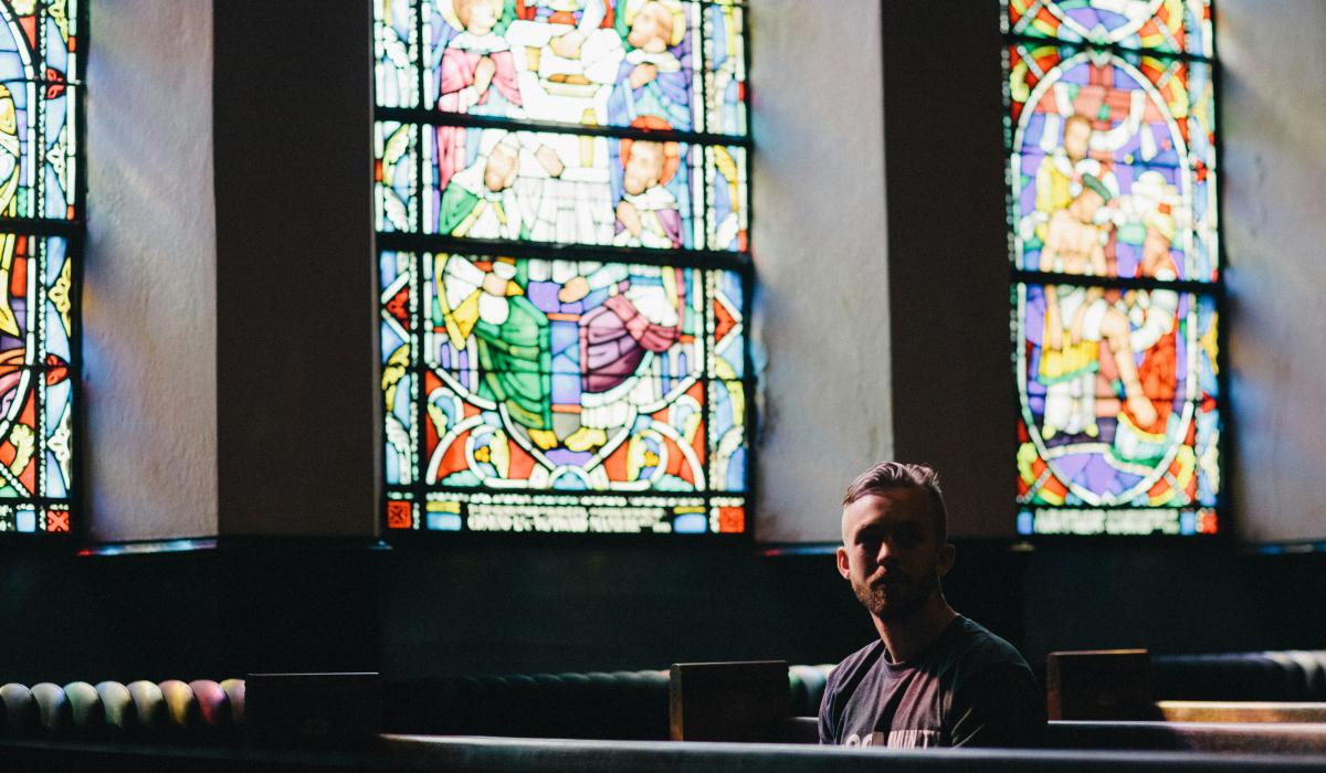 A man sits in a church pew below a colourful stained glass window, looking pensive.