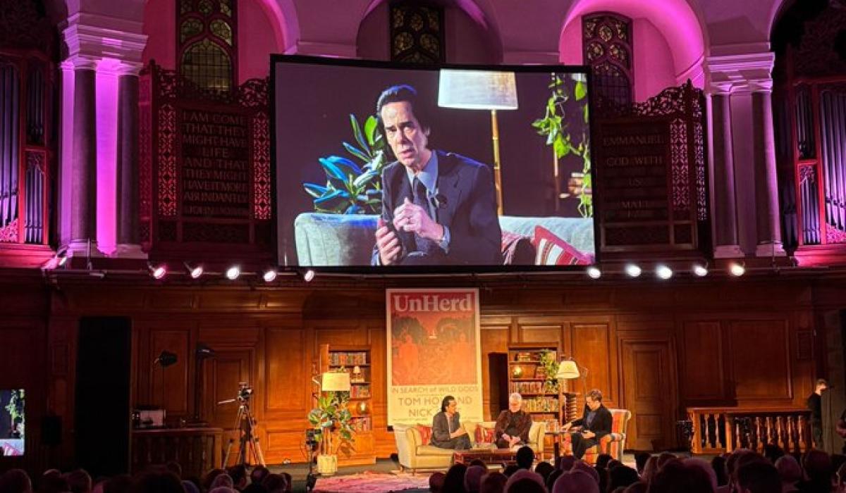 A view of an in-conversation on a stage, with a video screen above showing a close up.