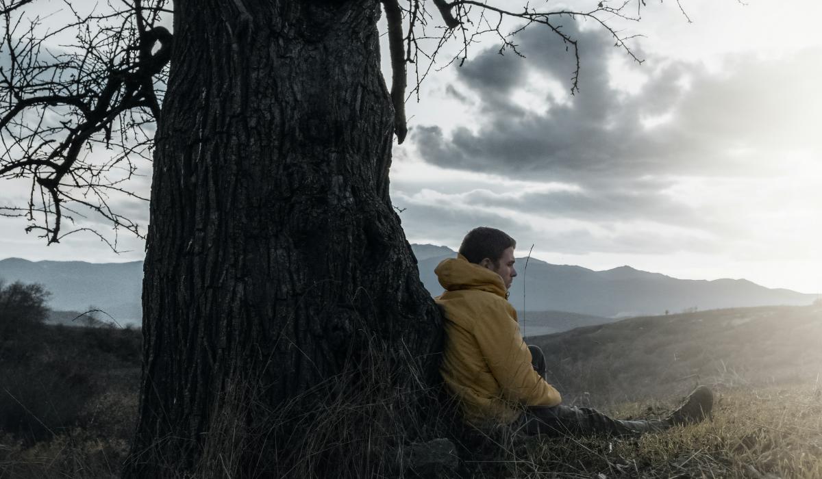 A wrapped-up man sits and leans against a bare tree, as dark clouds give way to sun.