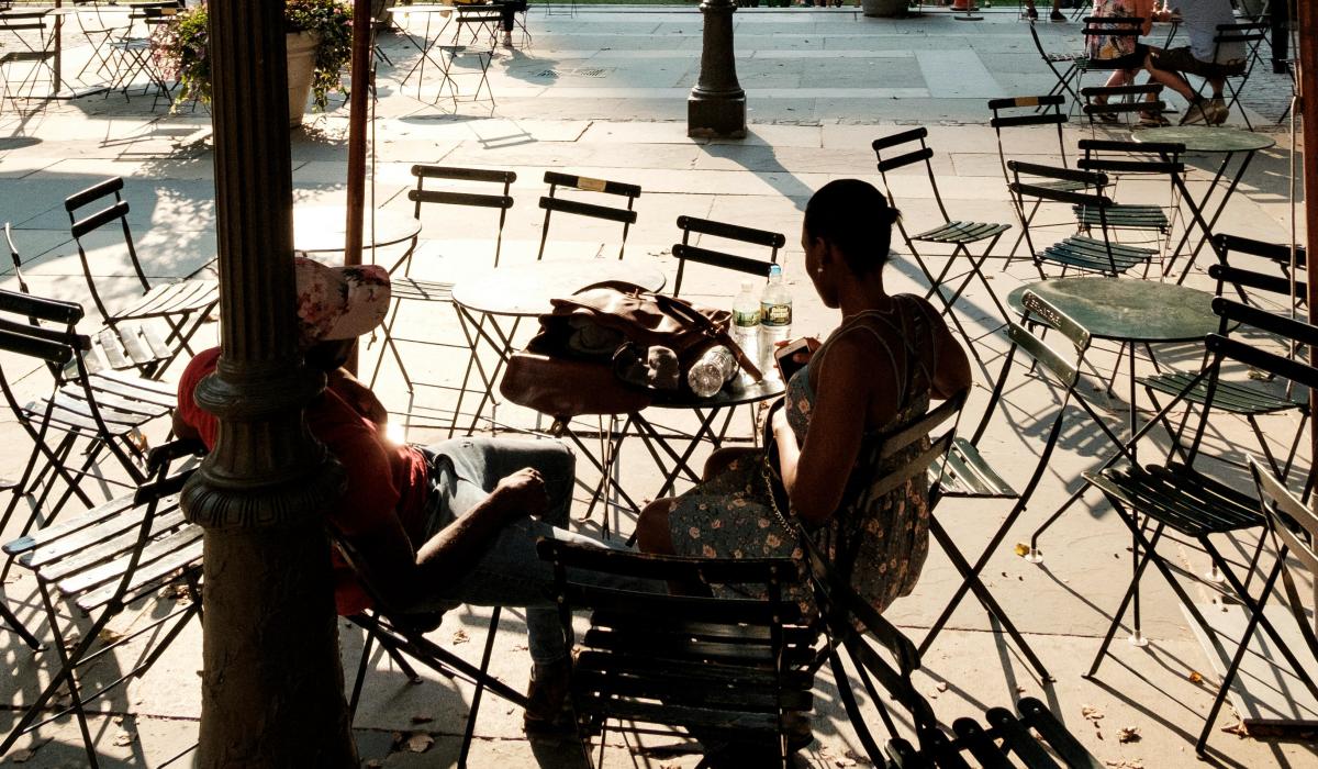 Two people. sitting at a street cafe amid empty tables and chairs, are silhoutted.