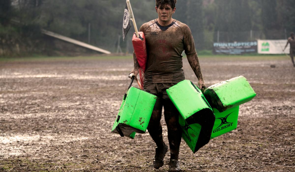 A muddy rugby player carries equipment off a pitch.