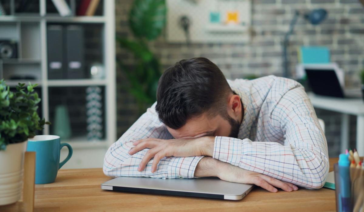 A man lies asleep on a closed lap top on a desk.