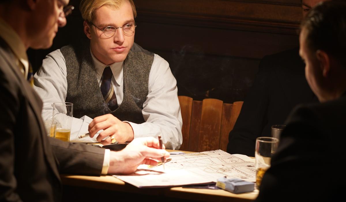 A man dressed in 1930s clothing, sits with others at a table looking pensive.