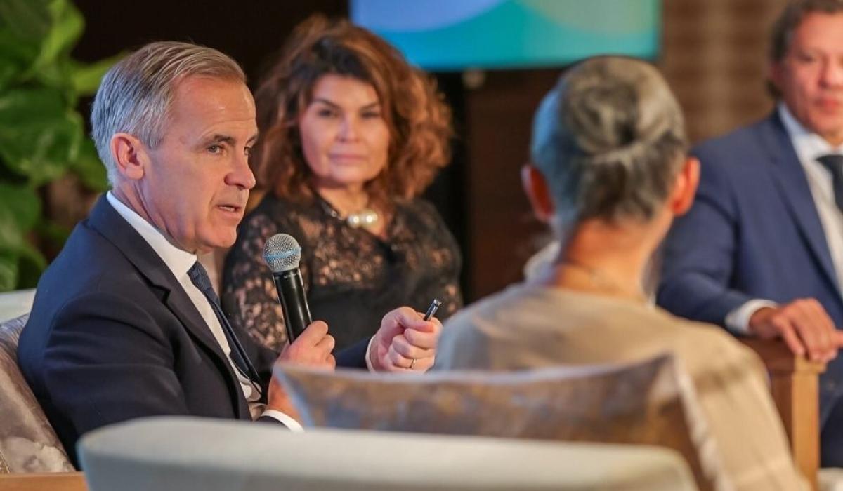 Mark Carney sits between two other speakers, holding a mic.